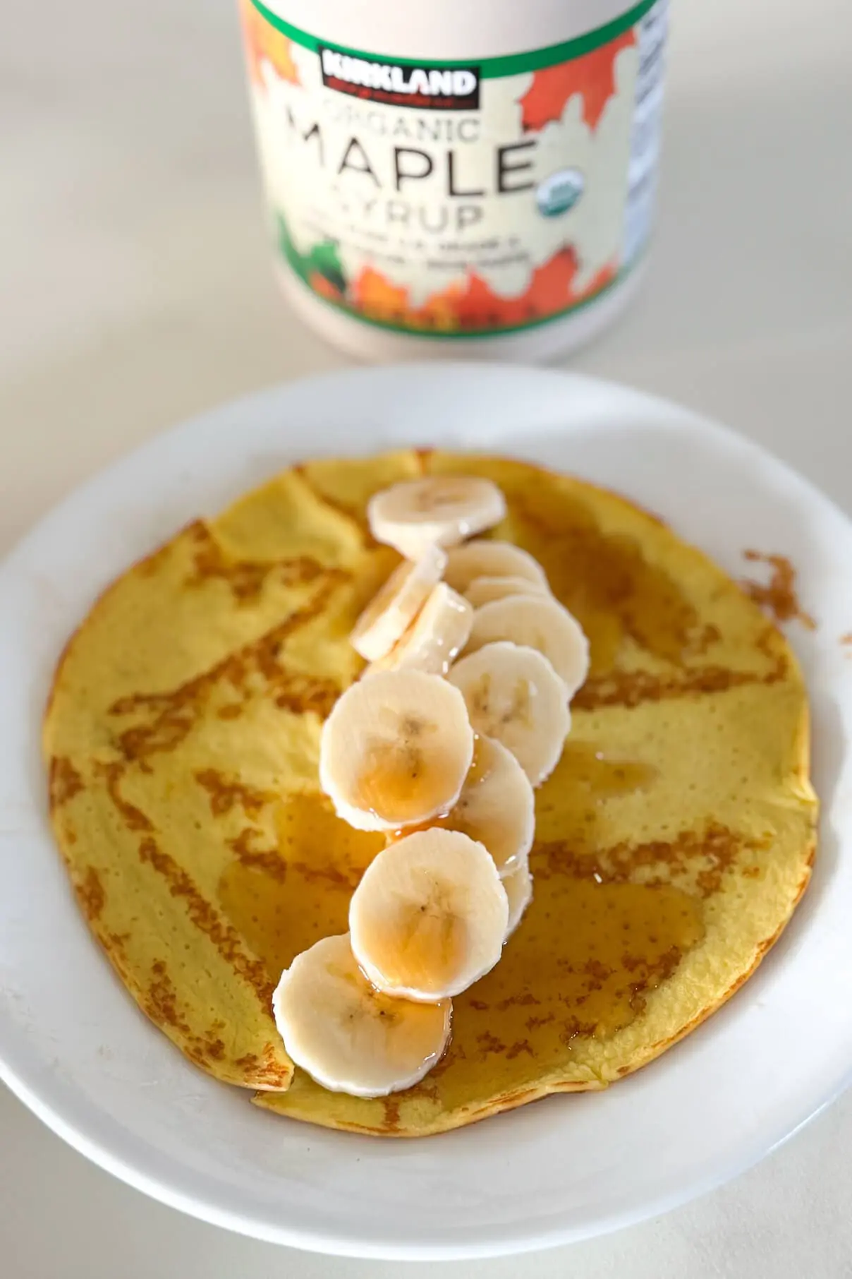 A plated pancake made with cottage cheese and eggs. It is garnished with fresh banana slices and maple syrup, styled with an organic syrup bottle slightly blurred in the background for depth.